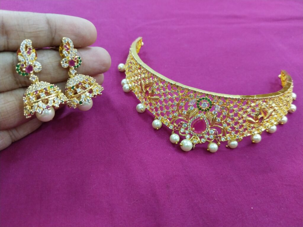 Gold choker with ruby and green center, pearl drops, and matching jhumka earrings on pink background