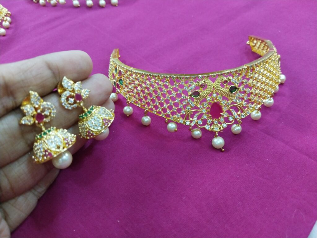 Gold mesh choker necklace with ruby stone and pearl drops, paired with matching jhumka earrings, displayed on pink fabric