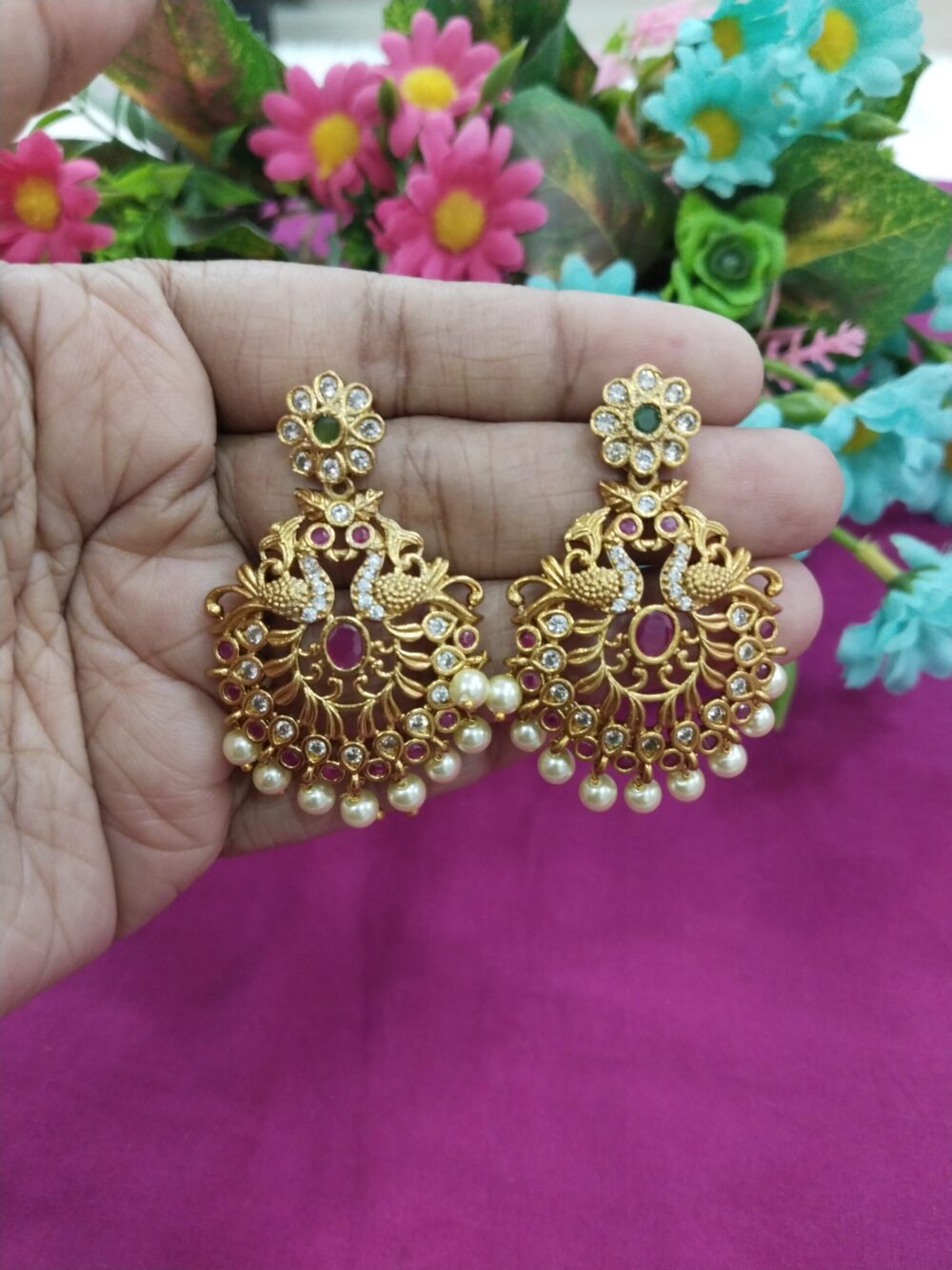 Gold-plated peacock design jhumka earrings with floral stud top, red stones, and pearl drops, held in hand with pink background.