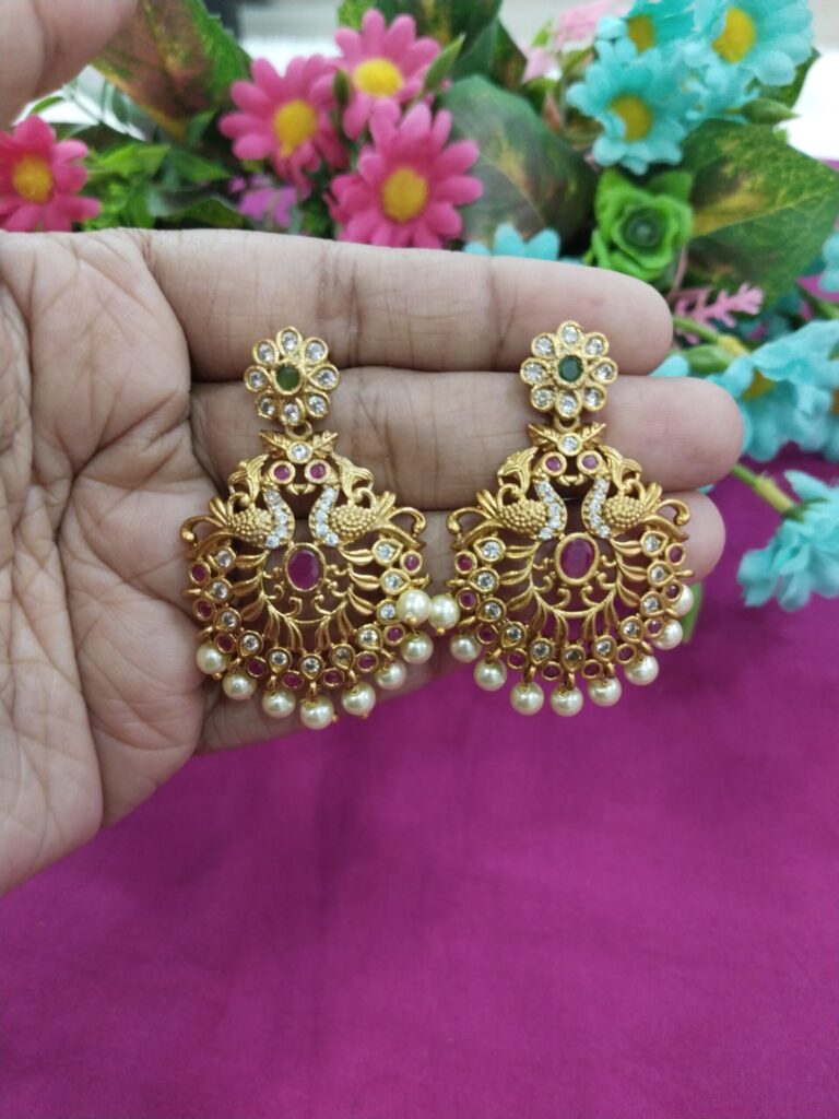 Gold-plated peacock design jhumka earrings with floral stud top, red stones, and pearl drops, held in hand with pink background.