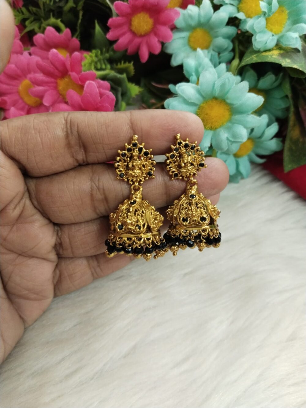 Close-up of gold Lakshmi temple jhumka earrings with black beads held against a floral background