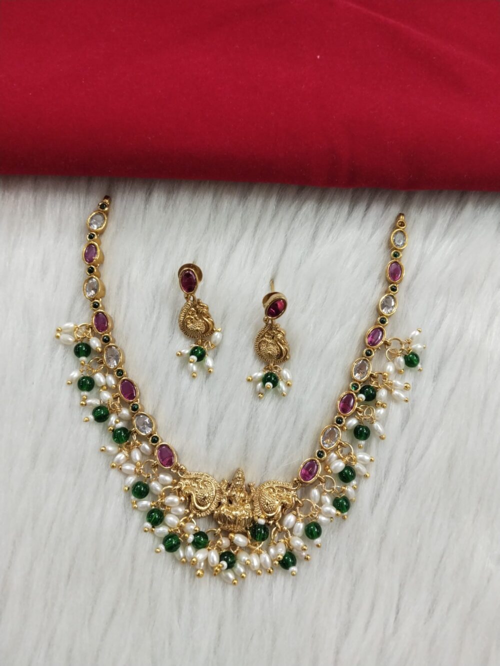 Traditional gold-plated temple jewelry necklace and matching earrings with ruby, emerald stones, and pearl accents on white fur background