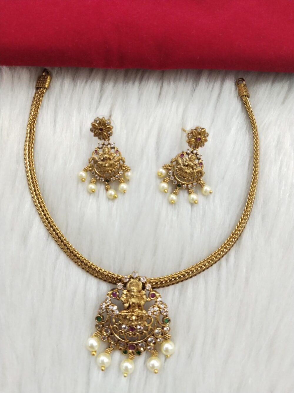 Antique gold-plated temple jewelry necklace set with Goddess Lakshmi pendant, colored stones, and pearl drops, placed on white fur background