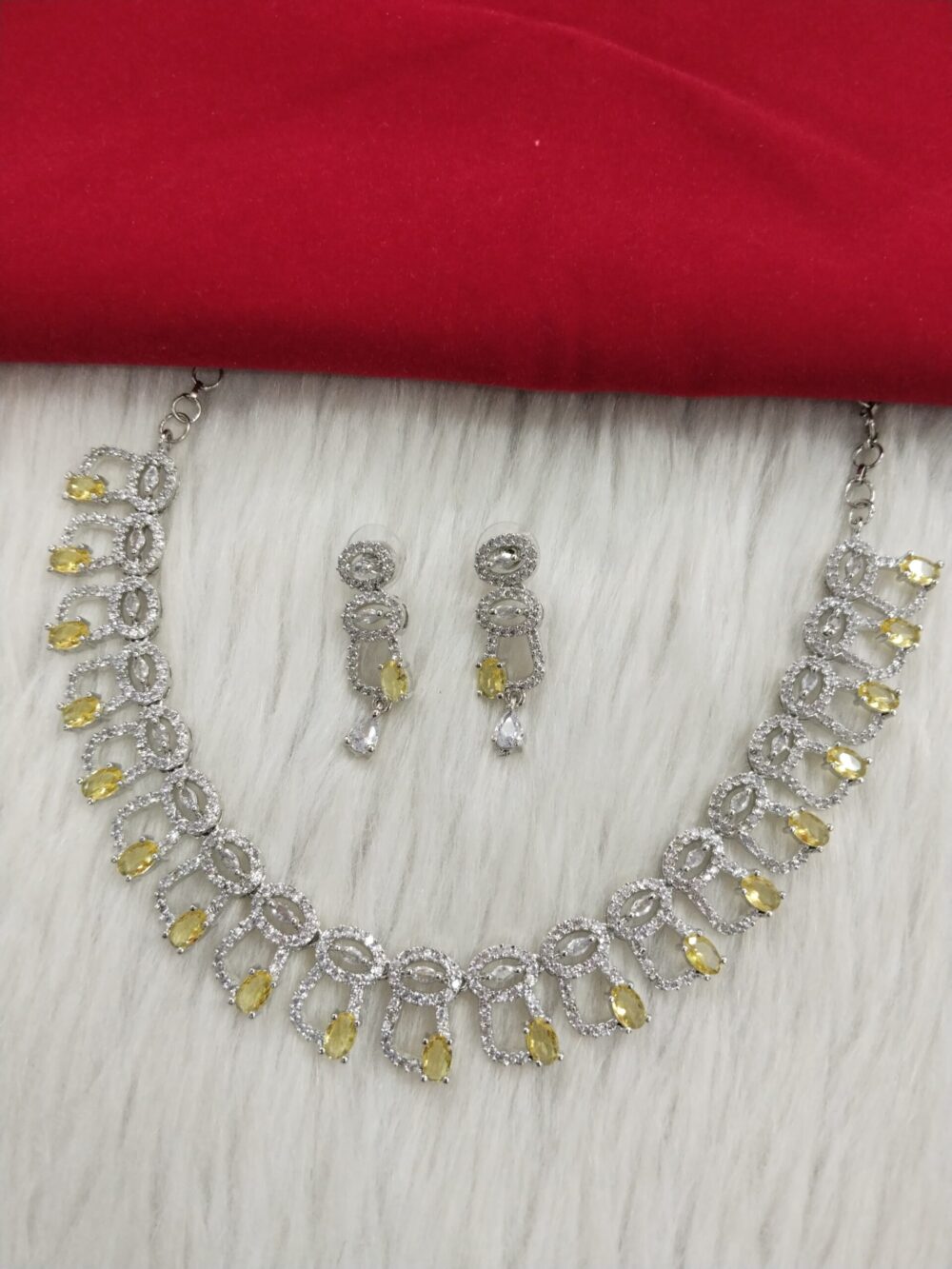 Silver finish necklace set with white American diamonds and yellow stone drops, paired with matching drop earrings, displayed on white fur with red fabric background.
