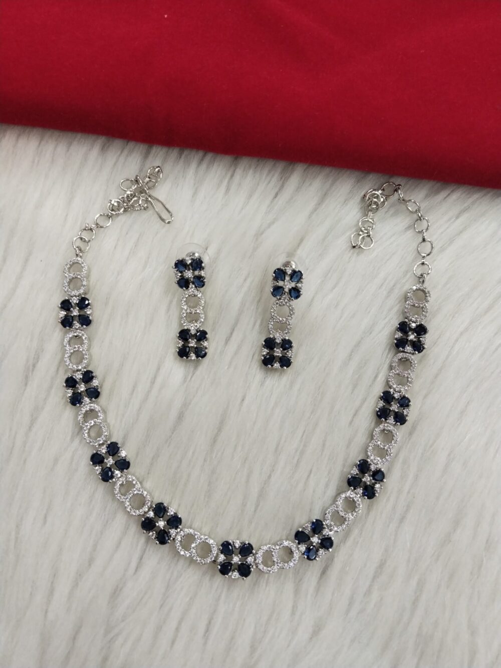 Silver necklace set with deep blue floral stones and white American diamonds, paired with matching earrings, displayed on a white fur surface and red cloth backdrop.