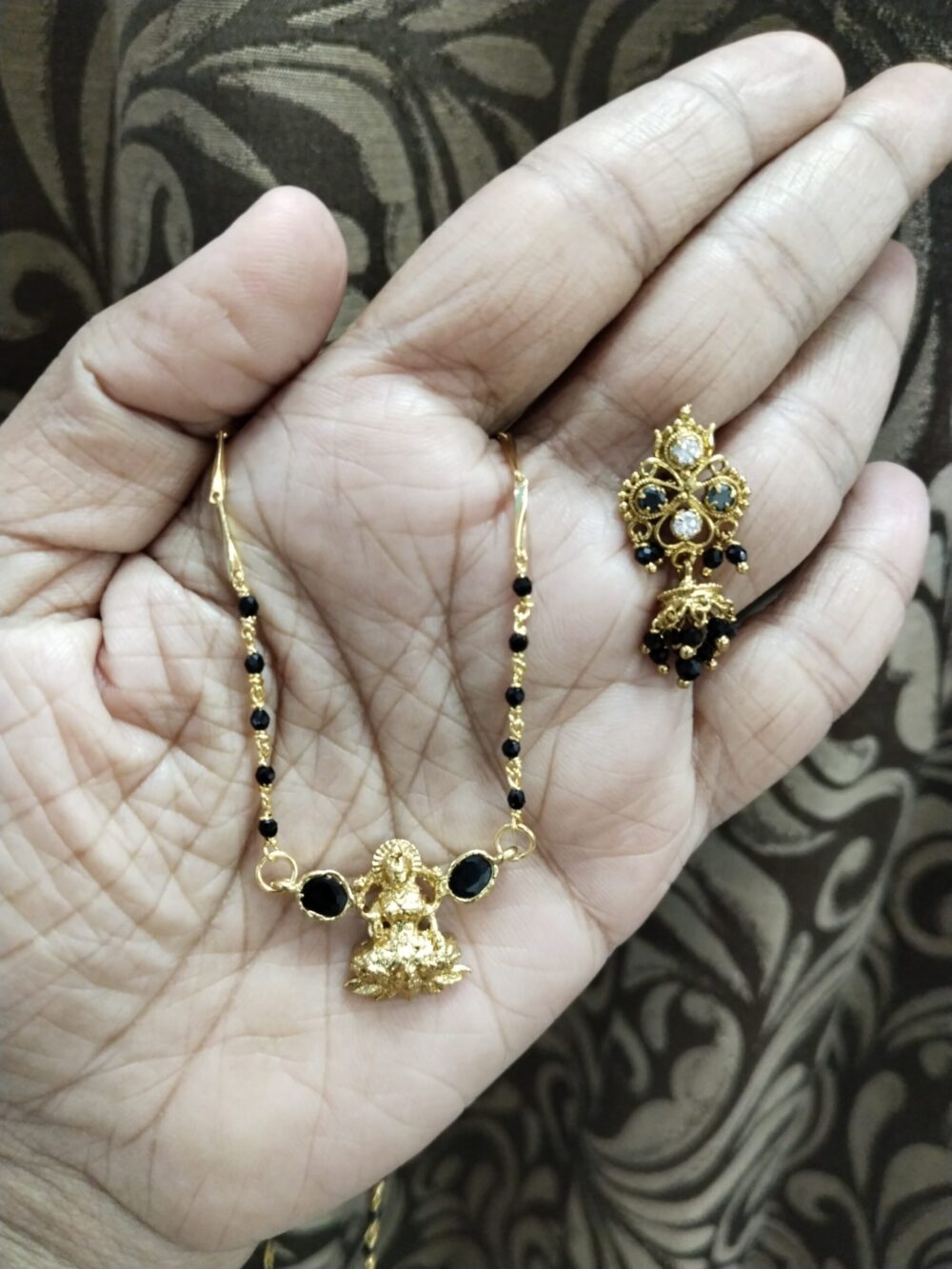 Hand holding gold-plated Lakshmi pendant mangalsutra with green CZ stones and matching multicolor stone ear cuff on traditional fabric background