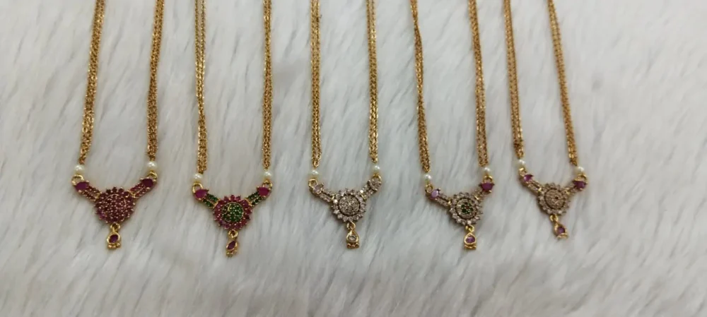 "Set of five traditional gold-plated chain necklaces with assorted pendant designs, featuring red, green, and white stones, pearl drops, and intricate detailing, displayed on a white fur background with a red fabric backdrop.