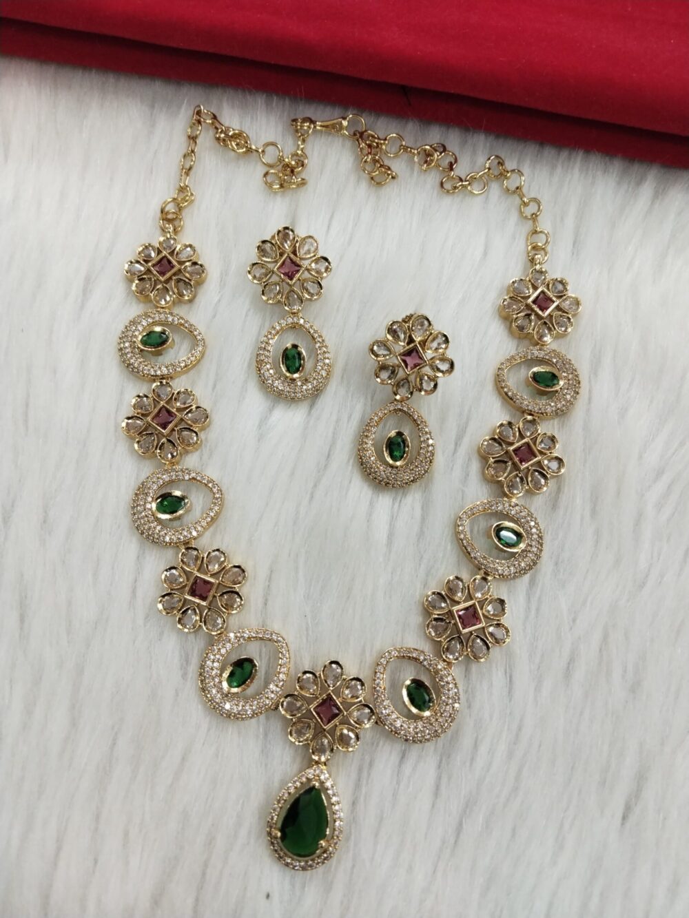 Gold-plated necklace and earrings set with green teardrop and ruby square stones on white fur background