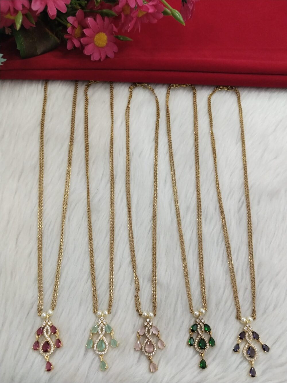 Set of 5 gold-plated pendant chains with red, green, pink, violet, and mint teardrop stone designs, arranged vertically on a white fur background with red cloth and flowers on top.