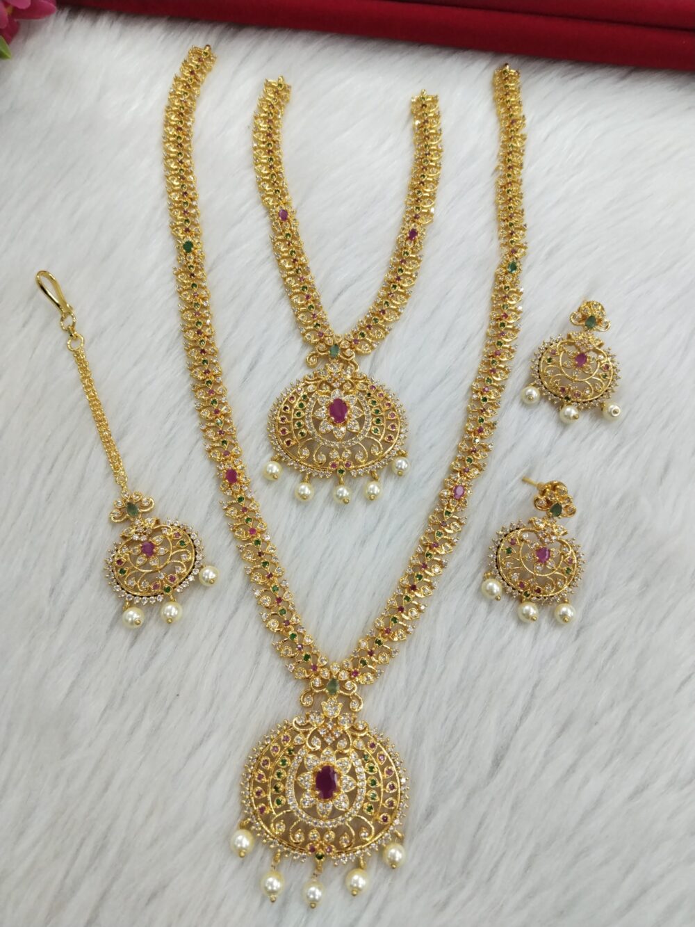 Traditional gold bridal jewelry set with round ruby pendants, green enamel accents, and pearl drops displayed on a white fur background